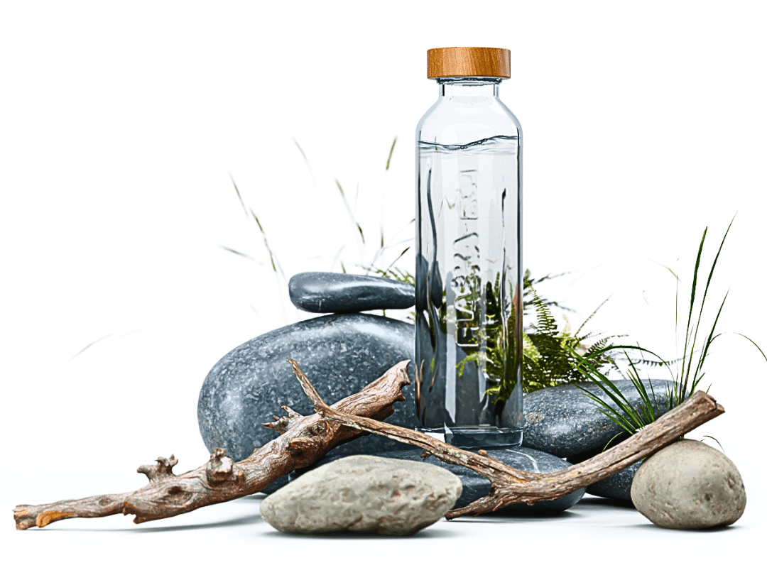 Glass water bottle with a bamboo lid standing among smooth gray stones, driftwood, and green grasses on a white background.